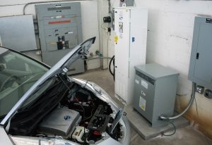 A Nissan Leaf electric vehicle has it's hood opened, charge cable plugged in, with three different charging units in the background of a laboratory, photo.