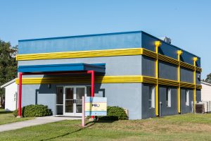 Small, one story commercial building painted in blue with yellow vents and red poles.