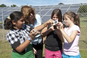 FSEC K-12 Education coordinator, Susan Scheith, evaluates three female students while they perform a hands-on activity with a hand-sized PV panel and fan motor with full-scale PV system in background behind a fence, photo.