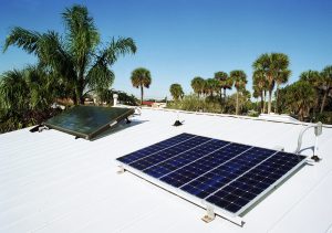 Solar water heating panel and solar electric panel on white metal roof of house with palm trees in background.