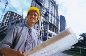 Man with yellow hardhat, holding blueprints, standing in front of tall commercial building under construction.