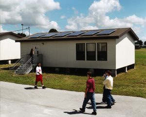 Portable classroom with solar electric panels on shingle roof with three students walking in front of while one student walks in opposite direction.