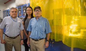 UCF researchers Ali Raissi, Nan Qin, Nazim Muradov stand alongside their solar-powered hydrogen production device, which is behind a transparent, yellow curtain in laboratory.