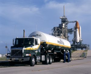 Air Products tanker of LH2 parked in front of Space Shuttle on launch pad at KSC. Two men in blue uniforms standing next to tanker.