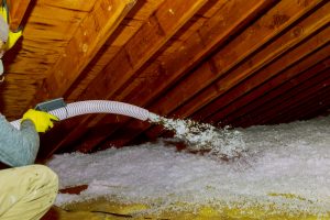 Technician spraying blown Fiberglass Insulation between Attic Trusses foam insulation repair tool in the white protect suit applies a construction foam from the gun to the roof.