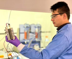 Asian male researcher holding fuel cell with equipment in laboratory hood, blurred in background.