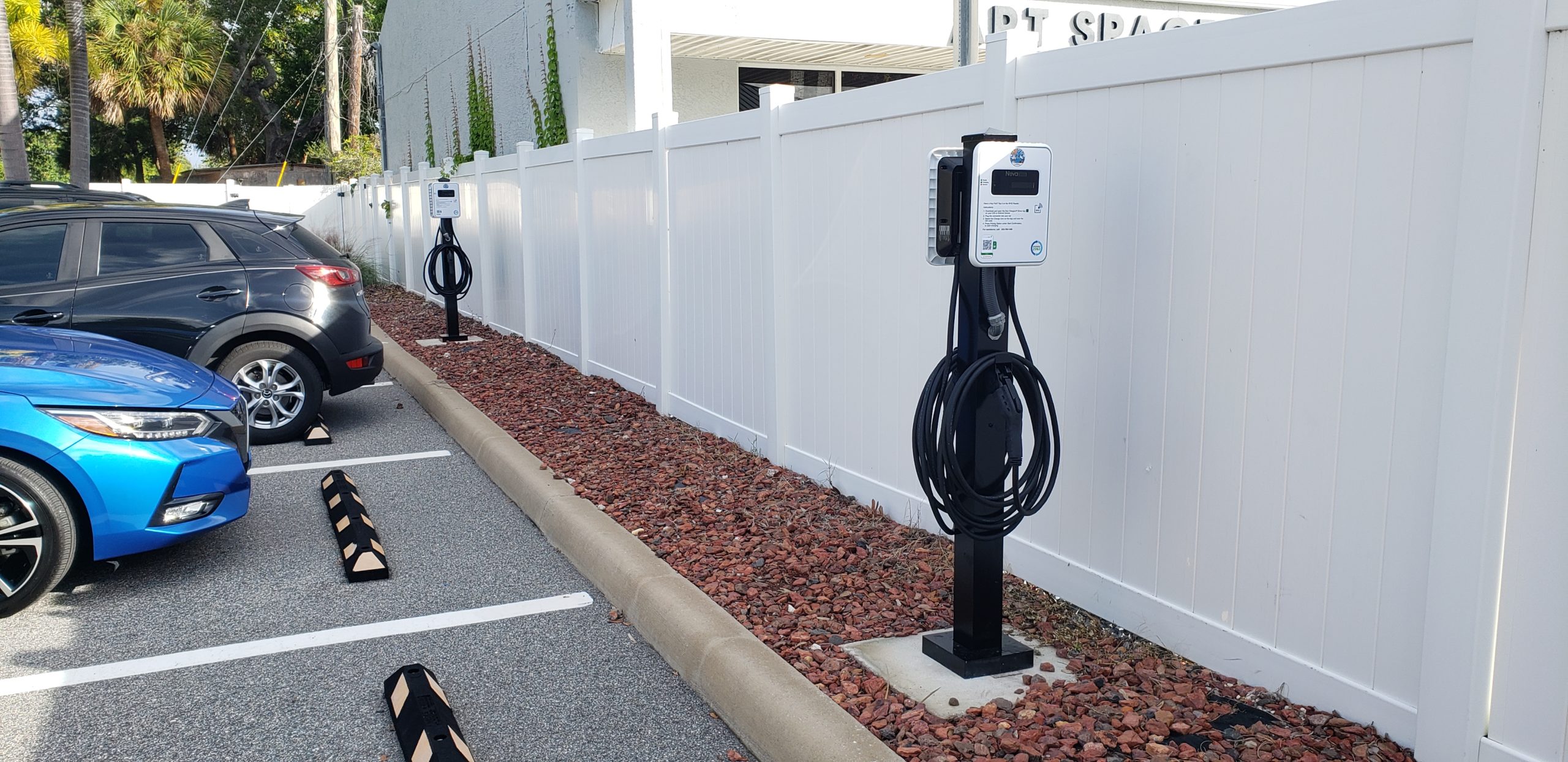 Two charging stations spaced apart with two parking spacing between them, with white plastic fence behind chargers. No cars are charging.